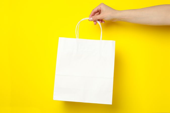 Female hand holding paper bag on yellow background, space for text