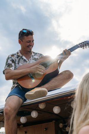 Man playing on guitar on car roof and women listening with pleasure