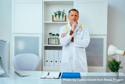 Contemplative mature doctor standing in a clinical setting with hand to his chin 5Xogv0