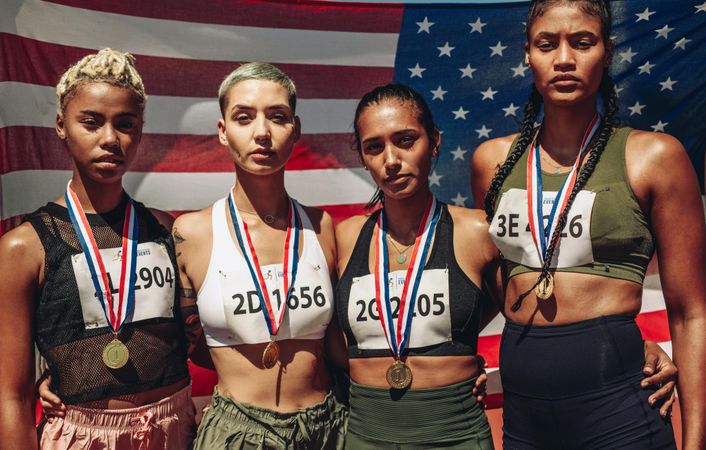 Portrait of four female athletes with gold medals standing in front of USA flag