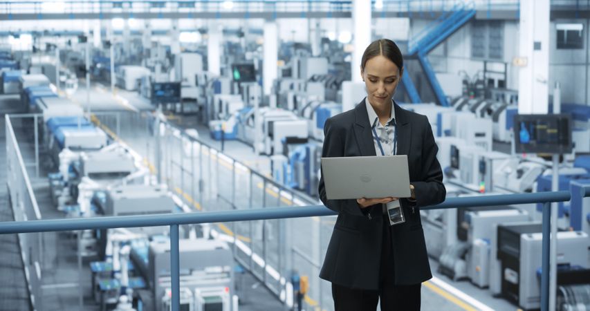 Portrait of a White Female Engineer Using Laptop Computer and Looking Around a Factory Facility with Robotic Arms Producing Modern Electronic Components for Tech Industries