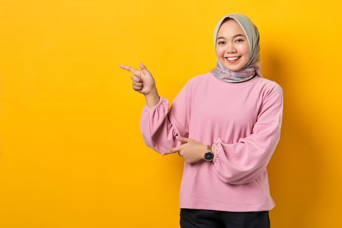 Happy Muslim woman in scarf and pink sweater gesturing towards copy space studio shoot