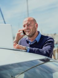 Vertical photo of a businessman talking on the phone while using a laptop supported by a car