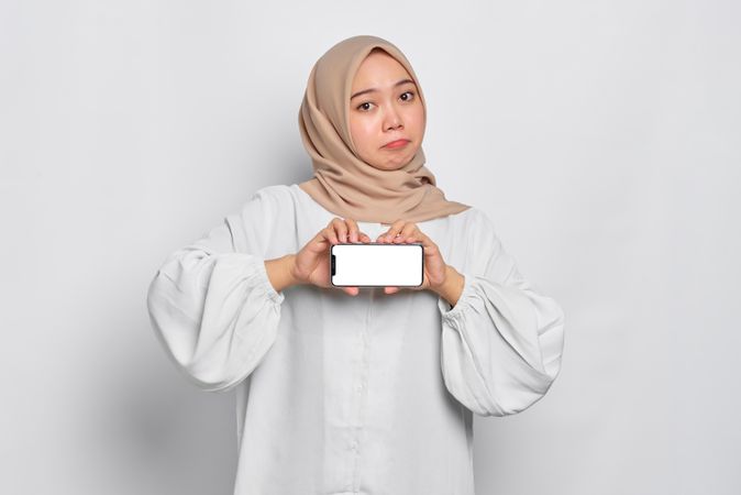 Questioning Asian Muslim woman in a bright studio shoot presenting cell phone with mockup screen
