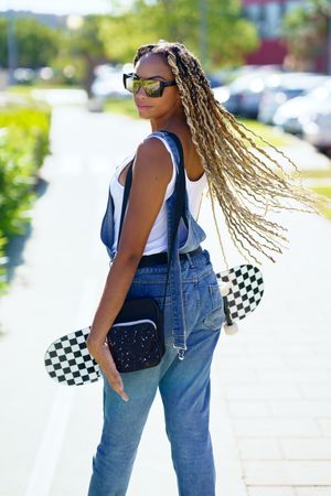 Female in denim overalls standing with skateboard and looking behind her