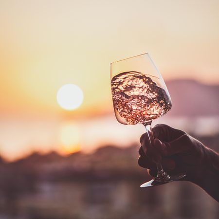 Man's hand holding glass of rose wine against sunset, square crop