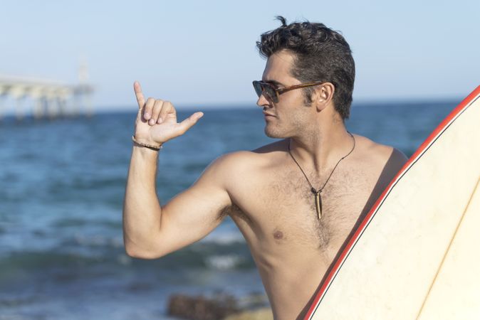 Male surfer standing with red outlined board near water making the “hang loose” sign