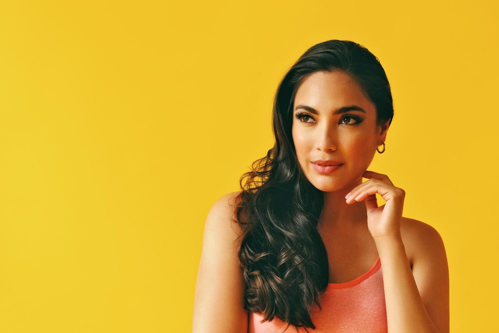 Headshot of calm Hispanic woman looking away in yellow room, with hand up to her cheek