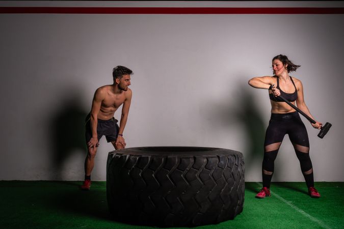 Man near woman with hammer hitting on tire in gym