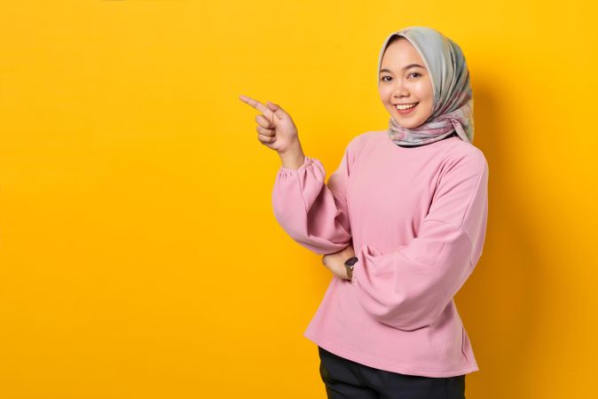 Smiling Muslim woman in scarf and pink sweater gesturing towards copy space studio shoot