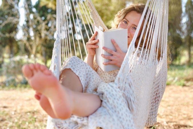 Cheerful woman on hammock looking at camera from behind book