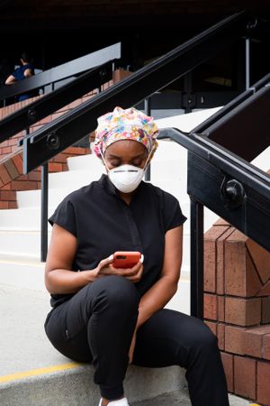 Nurse in head covering, N95 mask and black scrubs checks smart phone will on break