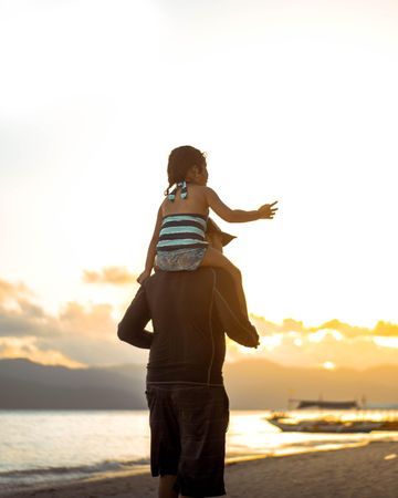 A man is carrying young daughter on his back standing on the beach at sunset