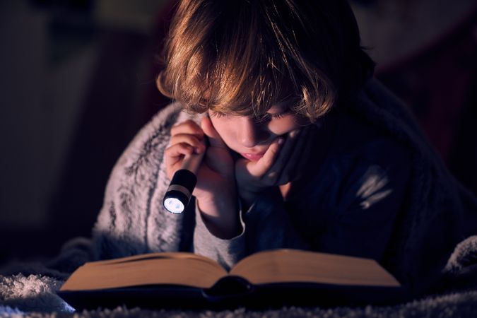 Focused boy reading book with flashlight