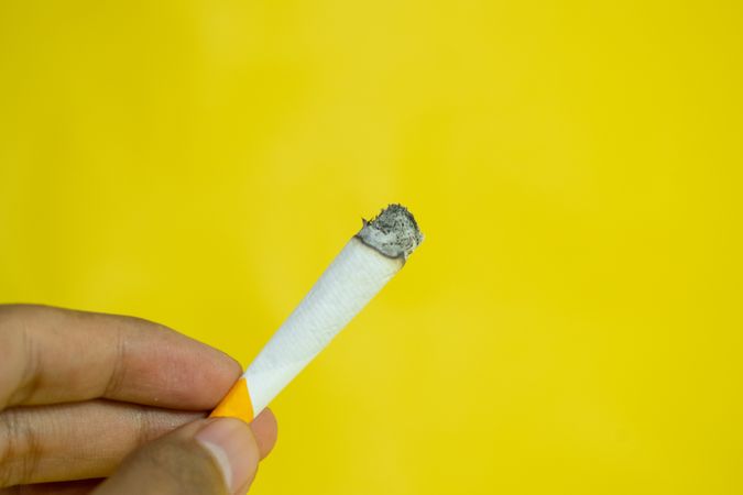 Yellow background with fingers holding lit hand rolled cigarette