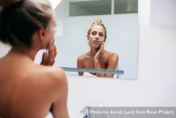 Beautiful young woman in bathroom looking in to mirror and applying moisturizer on face 5r3gM4