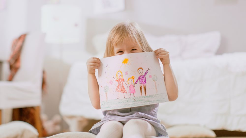 Young Girl Sitting on Pillows and Shows Drawing of Her Family and Hides Behind It.