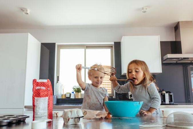 Young girl licking batter from spatula with her brother holding a whisk