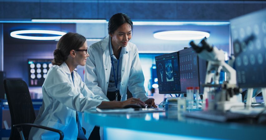 Asian And White Female Doctors Talking In Medical Research Center And Using Desktop Computer To Analyze Brain CT Scans.