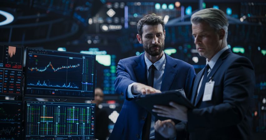 Team of Two Handsome Stock Exchange Brokers Using Tablet Computer, Chatting About the Asset Management Strategy for an International Business Client, Working in a Office Building with Computer Screens