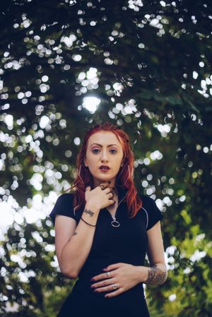 Young red-haired woman in makeup with tattoos looking at camera under tree with blurred background