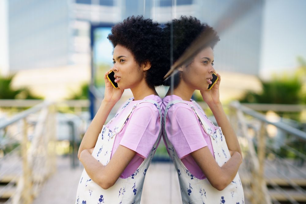 Female in floral overalls speaking on cell phone and leaning her back on window outside