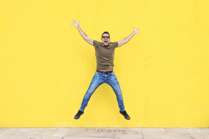 Excited male standing outside jumping in front of yellow wall with open arms