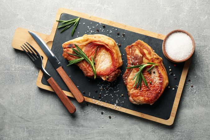 Board with steaks and cutlery on gray background. Grilled food
