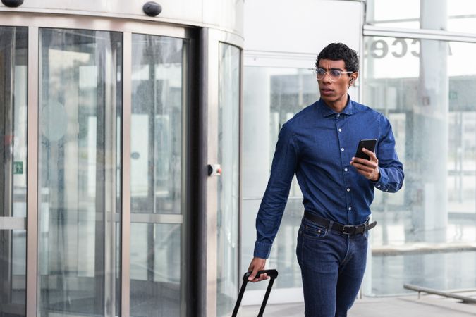 Stylish businessman with smartphone and luggage against glass wall