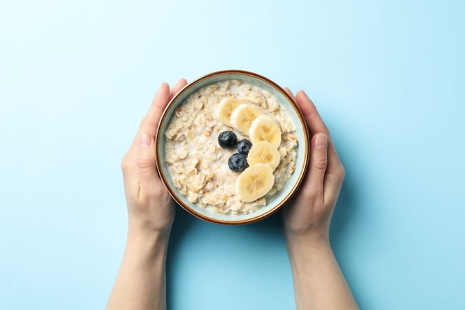 Female hands hold bowl with oatmeal porridge and fruits on blue background