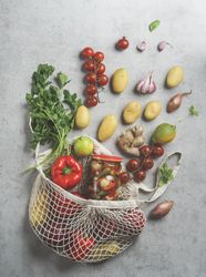 Sustainable groceries shopping bag with various vegetables: potatoes, tomatoes, pepper, lime, herbs, garlic, onion and preserve on grey concrete table