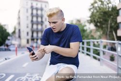 Young man with blonde hair sitting on the street surprised by something on a mobile phone 4OdL97