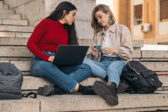 Two students sitting on the stairs of the college with a laptop. Watching mobile