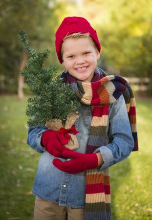 Young Boy Wearing Holiday Clothing Holding Small Christmas Tree Outside