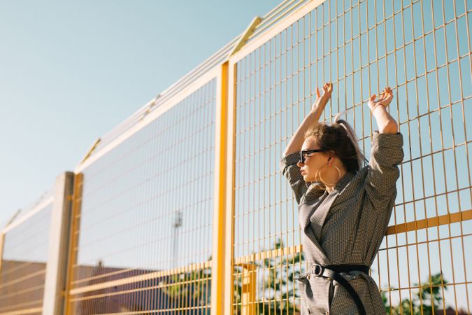 Stylish woman in sunglasses raising hands and leaning on lattice