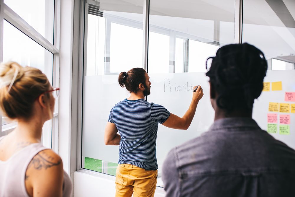 Man standing and writing at whiteboard with colleagues looking on