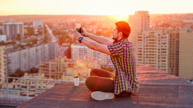 Male sitting on roof taking picture with tablet at dusk