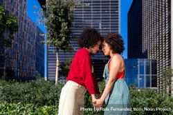 Female couple holding hands and facing each other in city park with office buildings in background 0KvWyb