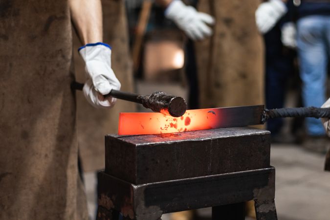 Blacksmith in gloves hot hitting metal part on anvil