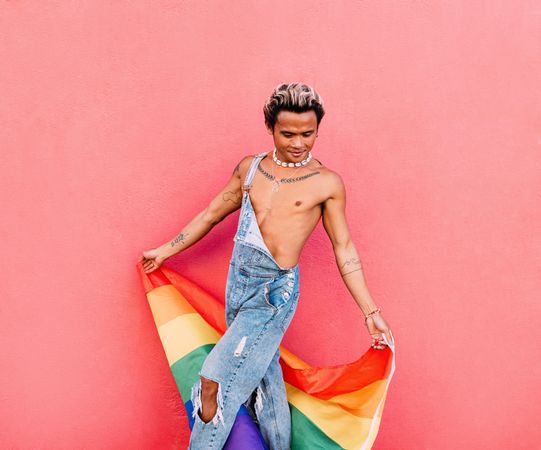 Male in open overalls looking down and holding a pride flag while leaning against a pink wall