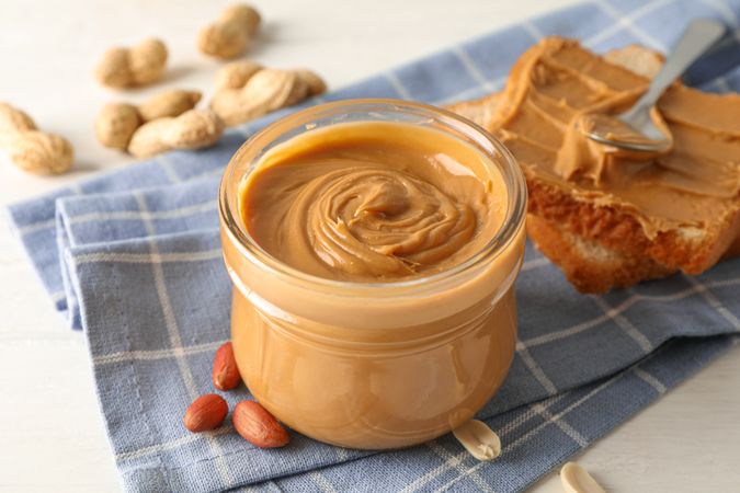 Glass jar with peanut butter, peanut, kitchen towel, spoon and peanut butter sandwich on wooden background, space for text and closeup