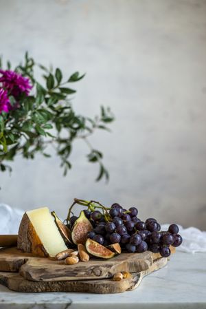 Ripe figs and dark blue grapes next to piece of cheese on cutting boards near pink flowers