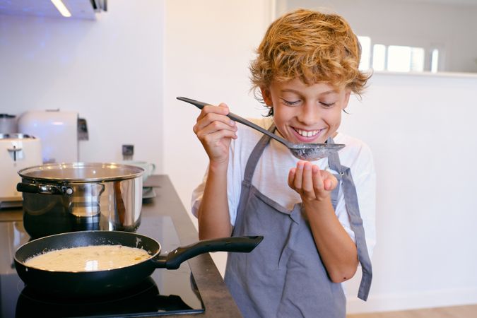 Cheerful boy with sauce in spoon cooking in kitchen