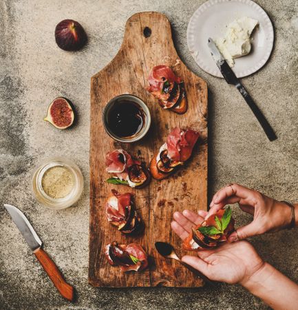 Hands holding crostini with prosciutto, goat cheese and grilled figs over wooden board