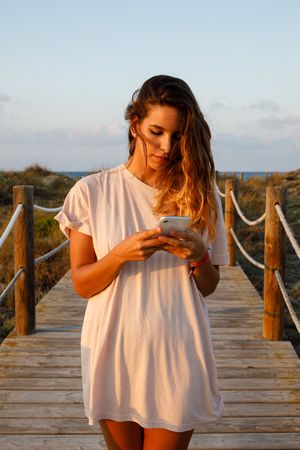 Tanned woman in white shirt walking on bridge and browsing smartphone