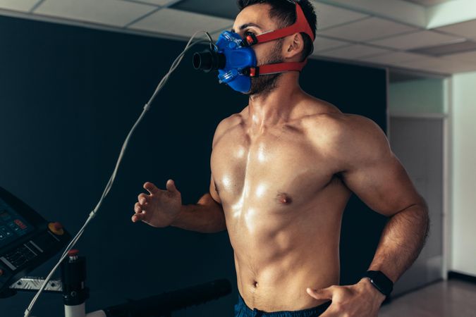 Muscular athlete with mask running on treadmill in gym for monitoring his performance
