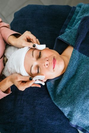 Crop beautician applying toner on face of female client in beauty salon