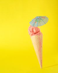 Cone with pink scoop of ice cream with cocktail parasol on yellow background