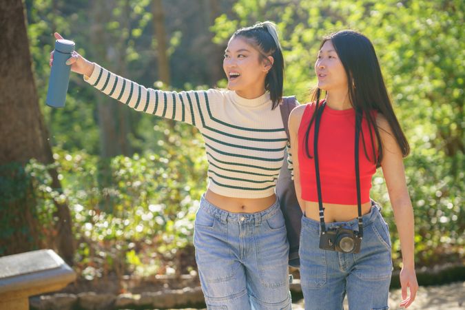 Asian female friends in casual apparel, standing in green forest of Alhambra and pointing at something in distance