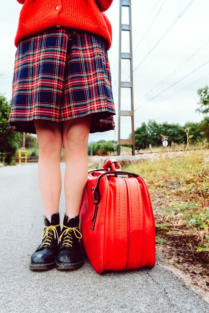 Woman in stylish boots with red suitcase standing on road